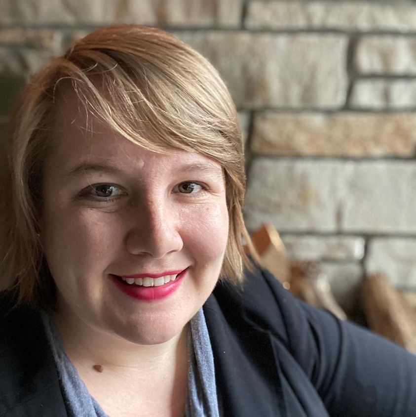 Stephanie Kunkel, a short haired blond woman, sits in front of a stone wall and firewood stack.  She wears a black blazer with a scoopneck grey shirt and bright red lipstick. 