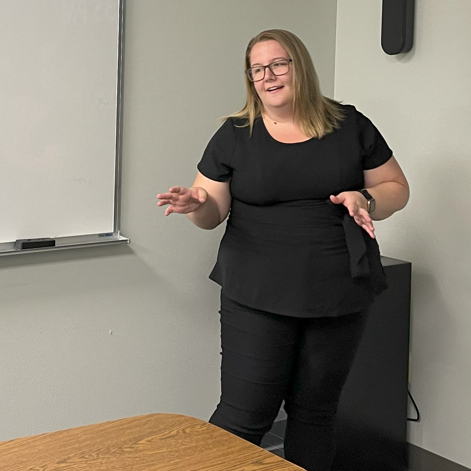 Stephanie Kunkel hosts a team building event in a board room, she stands in the corner near a white board she's dressed in black, her hands spread at her waist as she obviously discusses an important topic with the leaders in the room, she has blonde, shoulder length hair, dark glasses and a slight smile. She continues to shift perspectives.