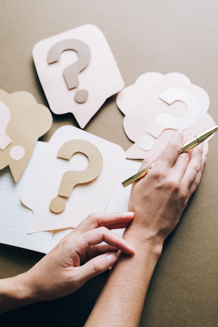 A person’s hands are resting on a table full of question marks. The person holds a metalic pen in their right hand as if they’re about to write, but need to think a bit more.