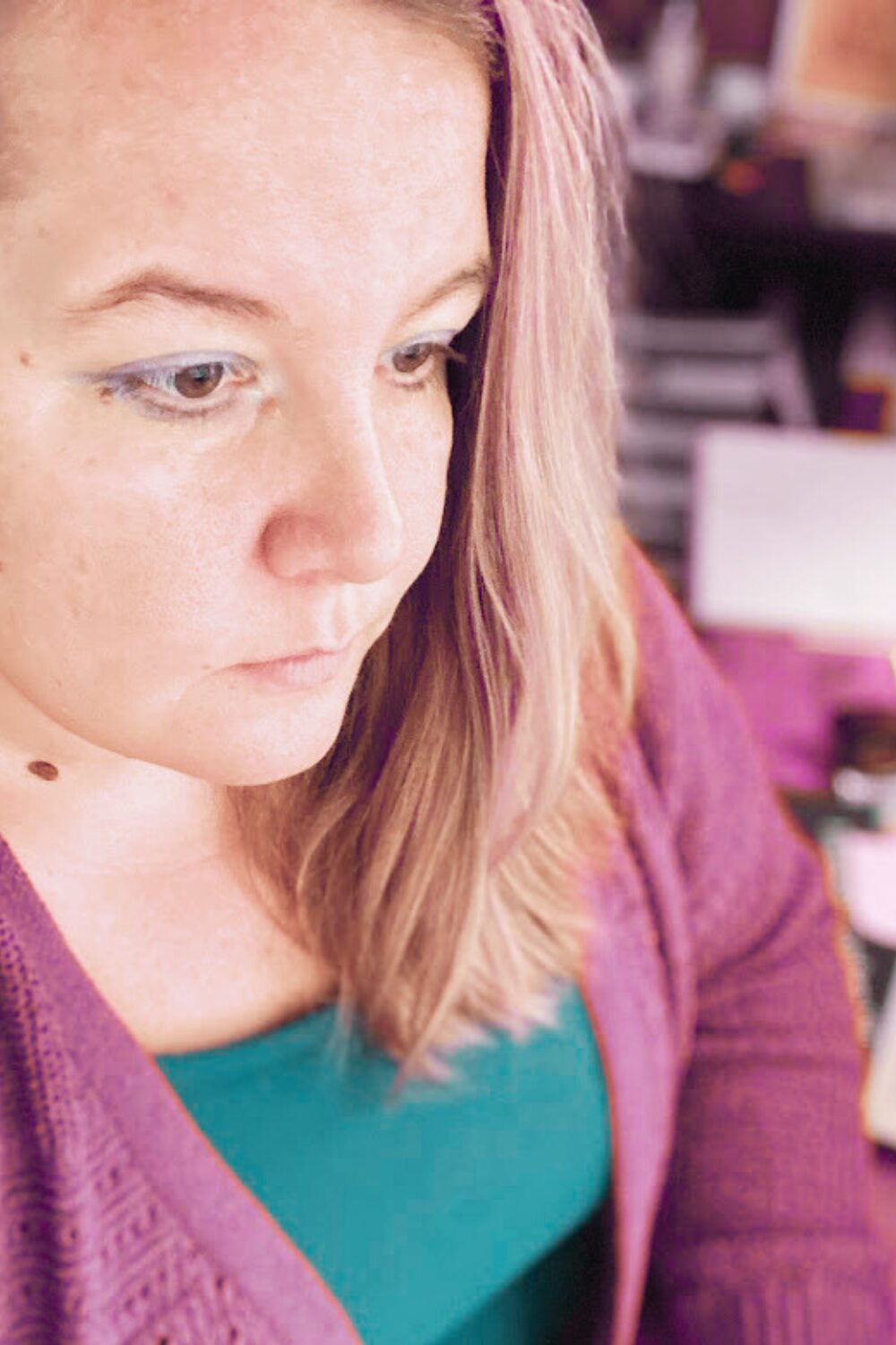 Stephanie Kunkel sits at her desk, working. She's wearing a green shirt and purple cardigan, her hair is a light shade or rose in this picture.