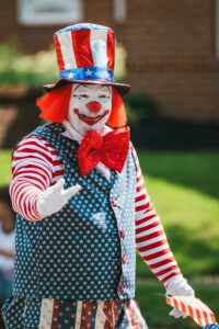 A clown dressed in red white and blue stars and stripes from head to toe.  He has a big red bow tie, red hair, a bright red nose and smile.  In his left hand he's holding a small american flag and his right hand is waving at someone outside of the frame.  
An American Clown.