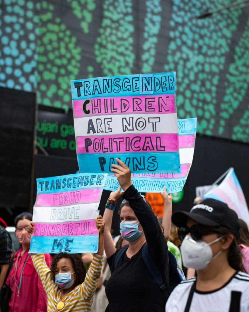 People at a protest hold signs advocating for transgender rights, including powerful messages like "Transgender children are not political pawns" and "Die for Trans People," emphasizing the urgency and importance of their cause.