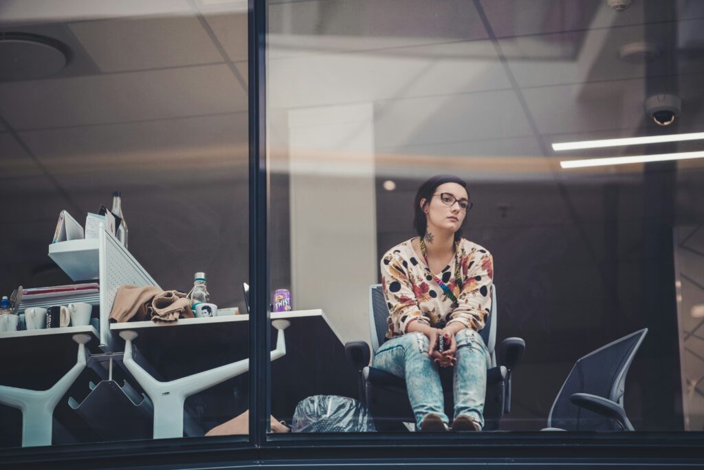 A woman in glasses, wearing a multicolored shirt and jeans, sits with her phone in her hands, and hands resting in her lap.  She stares out the window of what looks like her office, her feet propped against the window.  This relates to Stephanie describing how she is still operating like she is working in the corporate space.  The woman looks for more, and appears to be dreaming of better places.  She hasn't left the office yet. 